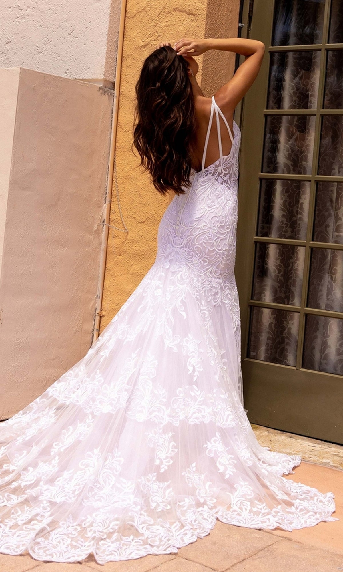 A woman in the Long Wedding Dress 11105 by Primavera, an ivory mermaid gown with lace embroidery and a long train, stands outdoors facing away near a glass door and beige wall.